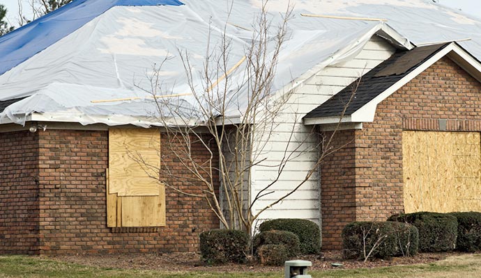 Storm-damaged brick house with boarded-up windows and a tarp covered roof